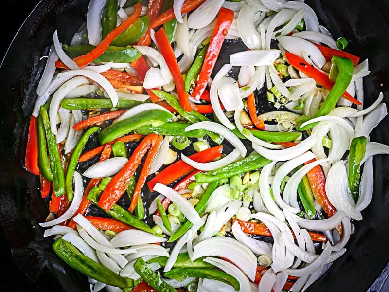 Image of sliced onions, bell peppers, Scotch bonnet pepper, green onion, garlic and fresh thyme being sautéed in a castiron skillet.