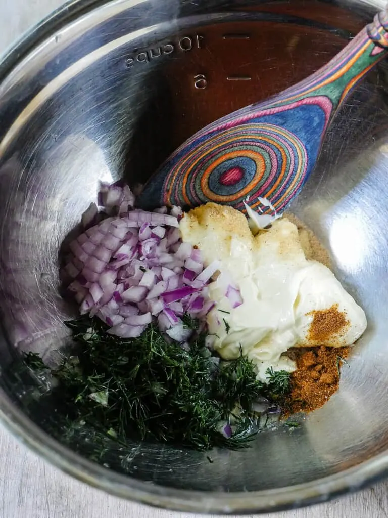 Image of a colorful spoon mixing mayonnaise, red onion, and fresh dill together in a metal mixing bowl.