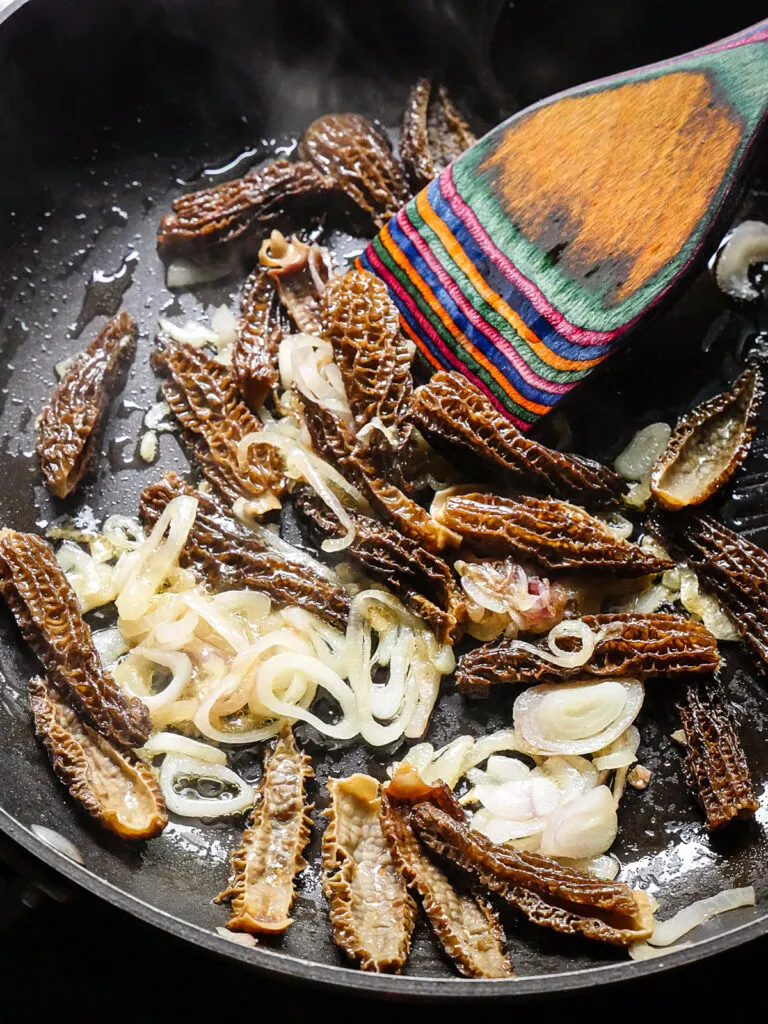 Image of shallots and morel mushrooms being sautéd in a skillet with a colorful wooden spoon.