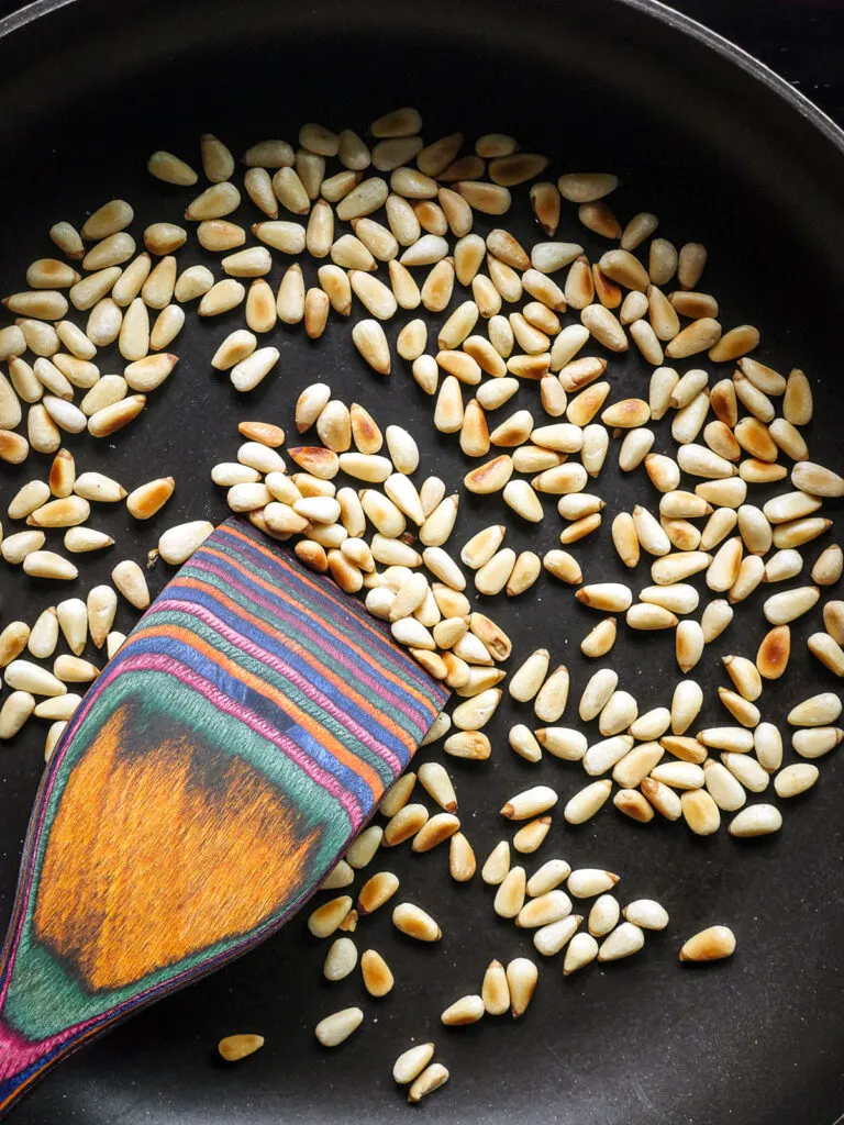 Close up of pine nuts being toasted in a skillet with a colorful wooden spoon.