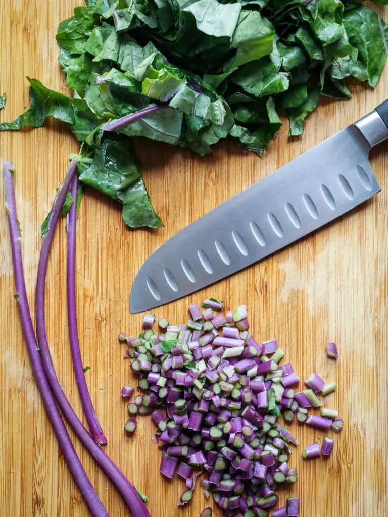 Separated kohlrabi greens and stems chopped on a wooden cutting board.