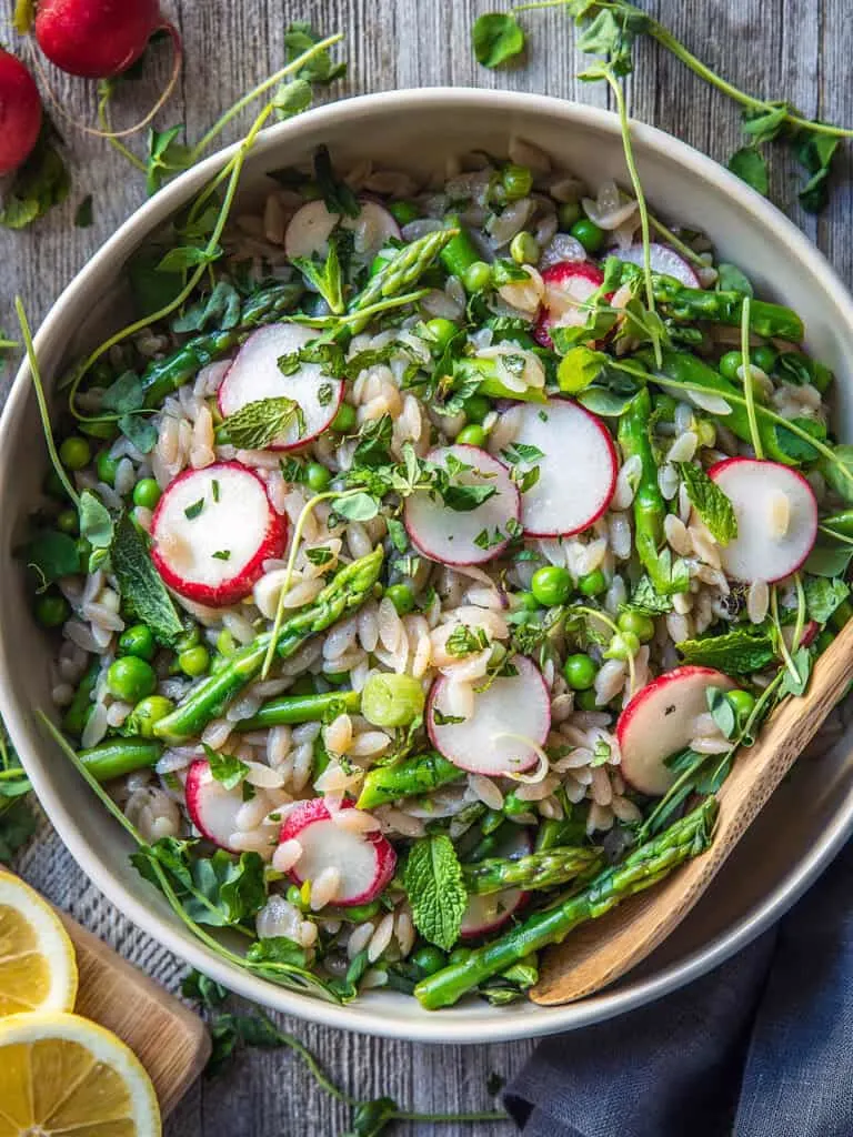 Styled shot of Cassava Orzo Salad with Asparagus, Peas, and Radishes in a white bowl on a wooden table.