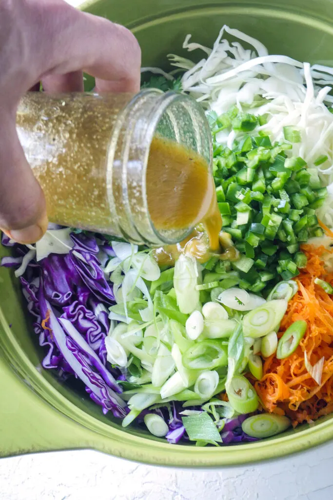 A hand pour dressing over a bowl of prepped coleslaw vegetables.
